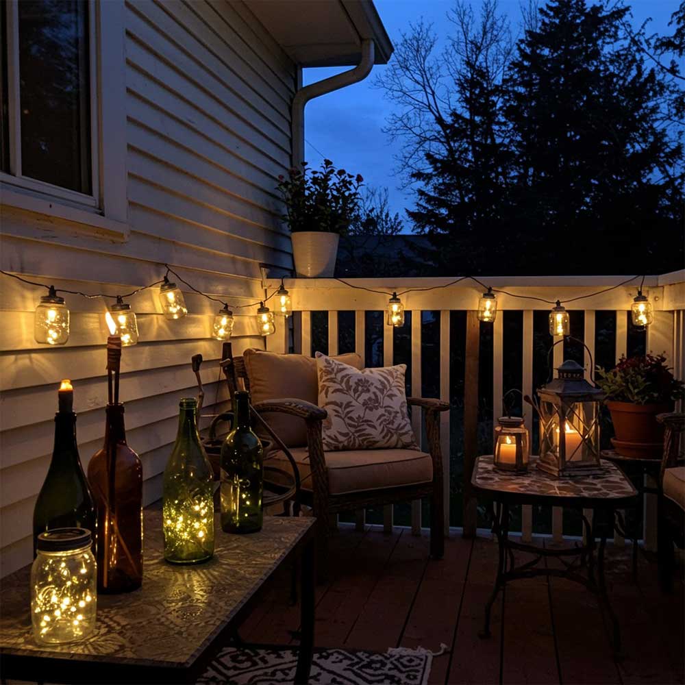 Small patio with budget lighting: mason jar lights, wine bottle torches, and reused lanterns, all glowing gently. Cozy decor on a tight budget.